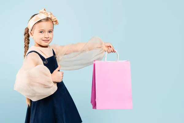 Happy kid with pigtails holding shopping bag and showing thumb up isolated on blue — Stock Photo