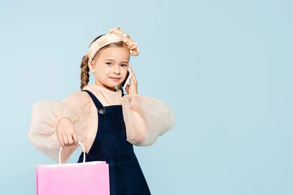 Happy kid with pigtails talking on smartphone and holding shopping bag isolated on blue — Stock Photo