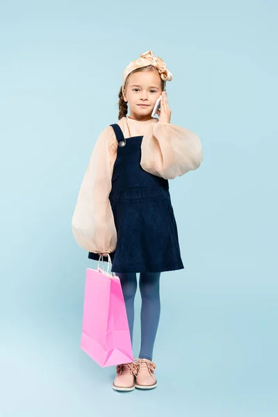 Full length of happy kid with pigtails talking on smartphone and holding shopping bag on blue — Stock Photo