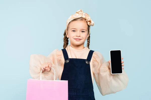 Glückliches Kind mit Zöpfen hält Smartphone mit leerem Bildschirm und Einkaufstasche isoliert auf blau — Stockfoto