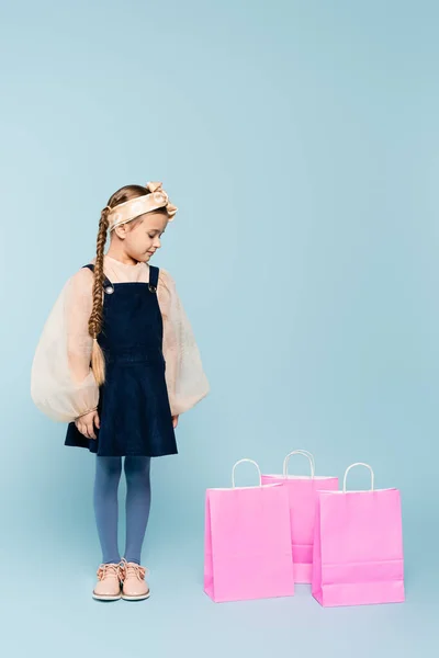 В полный рост маленькая девочка в платье смотрит на сумки с покупками синего цвета — стоковое фото