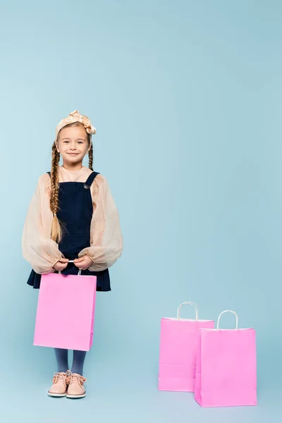 Longitud completa de niña en vestido sonriendo cerca de bolsas de compras en azul - foto de stock