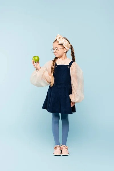 Pleine longueur de petite fille en robe et lunettes regardant pomme verte sur bleu — Photo de stock