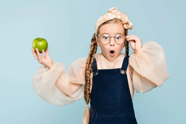 Шокована дитина регулює окуляри і тримає зелене яблуко ізольовано на синьому — стокове фото