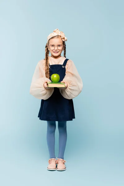 Piena lunghezza del bambino intelligente in bicchieri che tengono libro e mela verde su blu — Foto stock