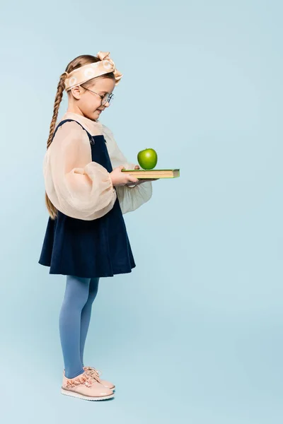 Longitud completa de niño inteligente en gafas mirando libro y manzana verde en azul — Stock Photo