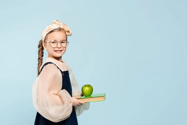 Bambino intelligente in bicchieri sorridente mentre tiene libro e mela verde isolato su blu — Foto stock