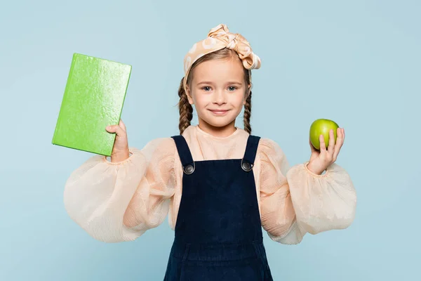 Niño inteligente sonriendo mientras sostiene libro y manzana verde aislado en azul - foto de stock