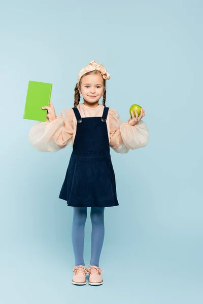 Full length of happy kid in dress holding book and green apple on blue — Stock Photo