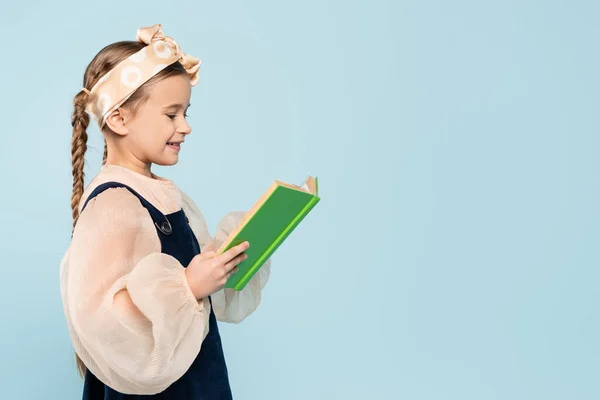Kleines Mädchen mit Zöpfen lächelt, während es Buch auf blauem Grund liest — Stockfoto