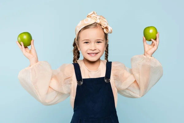 Little girl with pigtails smiling while holding green apples isolated on blue — Stock Photo