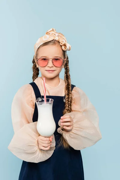 Маленькая девочка в солнечных очках, держащая стакан с молочным коктейлем, изолированным на голубом — стоковое фото