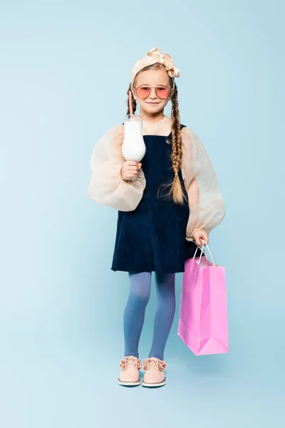 Longitud completa de niña en gafas de sol sosteniendo vidrio con batido y bolsa de compras en azul - foto de stock