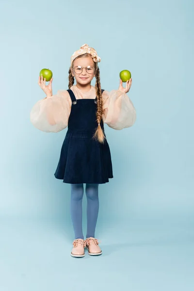 Pleine longueur de gosse dans des lunettes tenant des pommes vertes sur bleu — Photo de stock