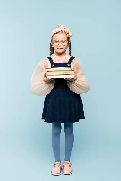 Повна довжина розумної дитини в окулярах посміхається, тримаючи книги на синьому — стокове фото