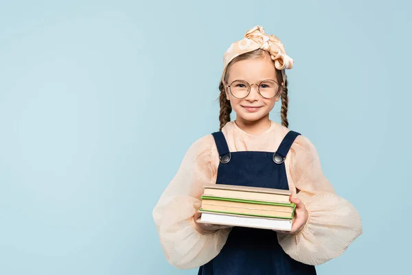 Garoto inteligente em óculos sorrindo enquanto segura livros isolados em azul — Fotografia de Stock