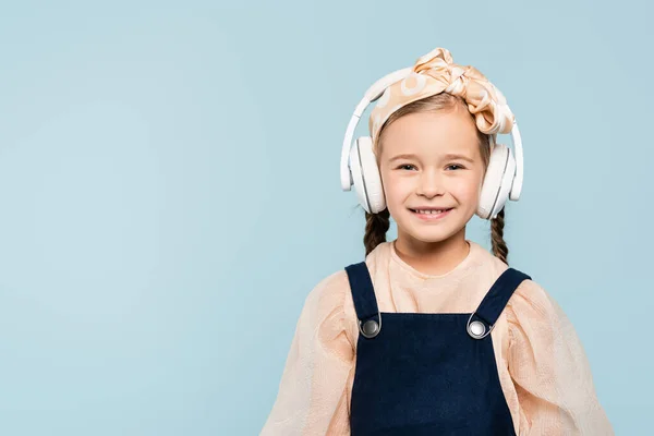 Fröhliches Kind im Stirnband mit Schleife und kabellosen Kopfhörern, das isoliert auf blauem Grund Musik hört — Stockfoto