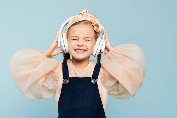 Garoto satisfeito em headband com arco e fones de ouvido sem fio ouvir música isolada no azul — Fotografia de Stock