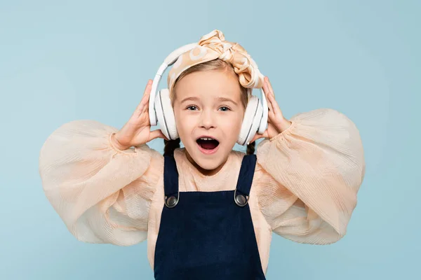 Amazed kid in headband with bow and wireless headphones listening music isolated on blue — Stock Photo