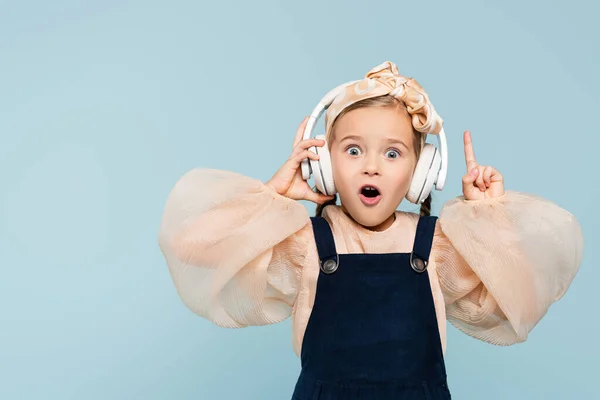 Chaval sorprendido en auriculares inalámbricos escuchando música y señalando con el dedo aislado en azul - foto de stock