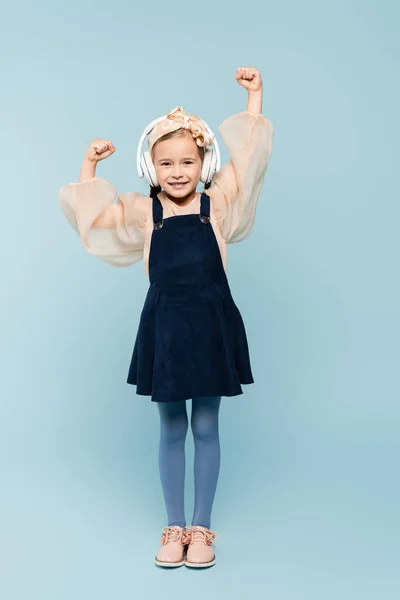 Full length of cheerful kid in headband with bow and wireless headphones listening music on blue — Stock Photo