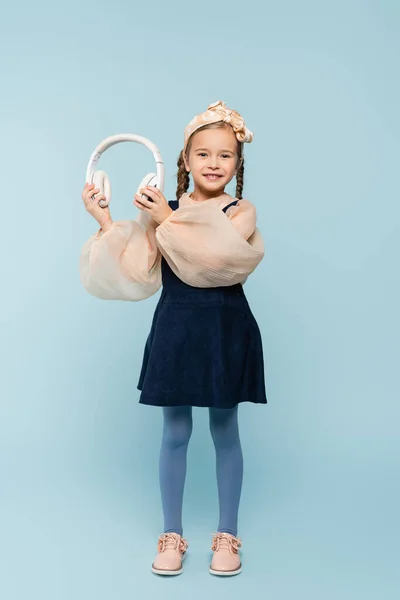 Full length of cheerful kid in headband with bow holding wireless headphones on blue — Stock Photo