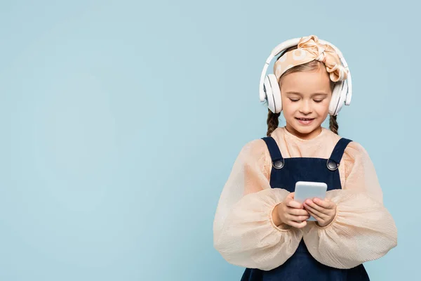Fröhliches Kind im Stirnband mit Schleife und drahtlosen Kopfhörern SMS auf Smartphone isoliert auf blau — Stockfoto