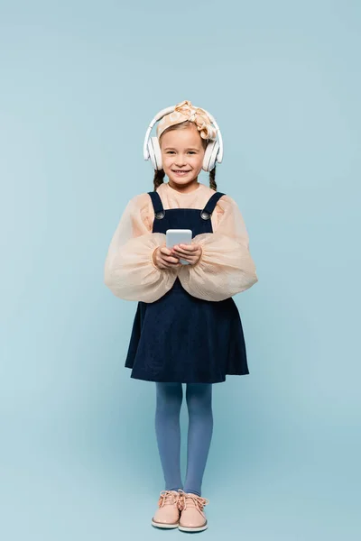 Full length of cheerful kid in headband with bow and wireless headphones using smartphone isolated on blue — Stock Photo