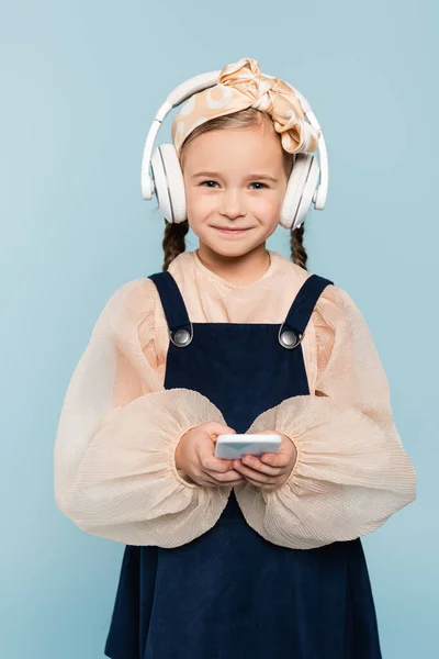 Cheerful kid in headband with bow and wireless headphones holding smartphone isolated on blue — Stock Photo