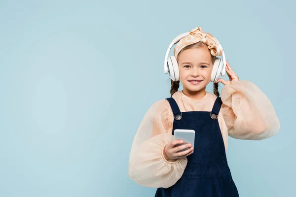 Cheerful kid in headband with bow adjusting wireless headphones while holding smartphone isolated on blue — Stock Photo