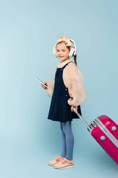 Longitud completa de la niña feliz en los auriculares inalámbricos utilizando el teléfono inteligente, mientras que de pie con el equipaje en azul - foto de stock