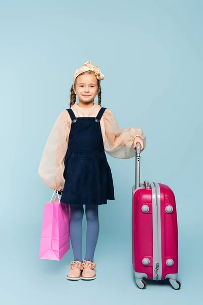 Pleine longueur de heureuse petite fille tenant sac à provisions et debout avec des bagages sur bleu — Photo de stock