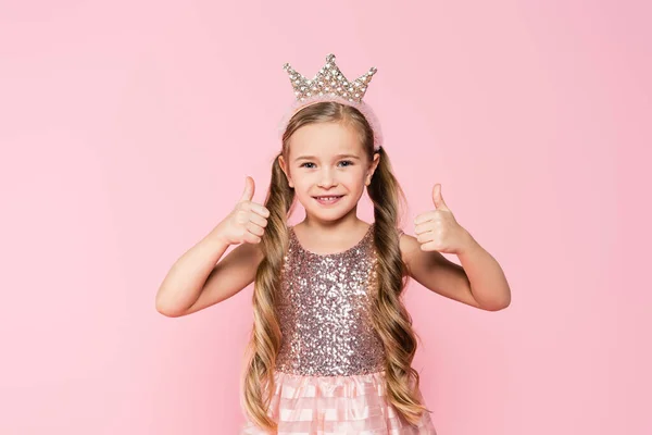 Alegre niña en vestido y corona mostrando los pulgares hacia arriba aislado en rosa - foto de stock