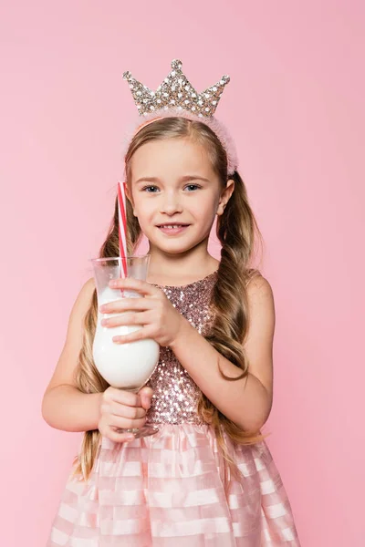 Joyeuse petite fille en robe et couronne tenant verre avec milkshake isolé sur rose — Photo de stock