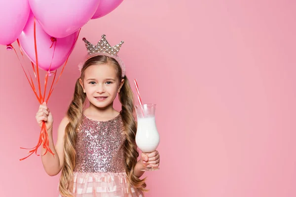 Joyeuse petite fille en robe et couronne tenant verre avec milkshake et ballons isolés sur rose — Photo de stock