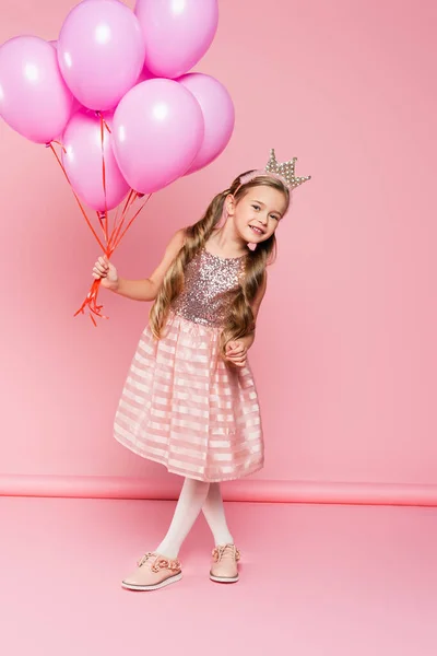 Pleine longueur de joyeuse petite fille en robe et couronne tenant des ballons sur rose — Photo de stock