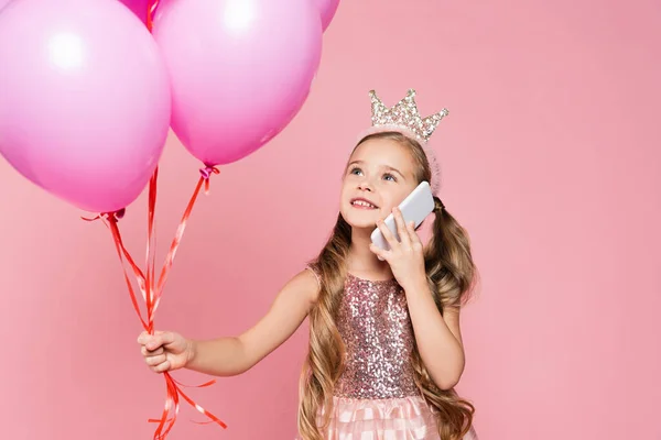 Niña alegre en vestido y corona sosteniendo globos y hablando en teléfono inteligente aislado en rosa - foto de stock