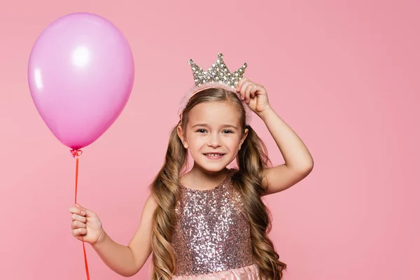 Gioiosa bambina in abito regolazione corona e tenendo palloncino isolato su rosa — Foto stock
