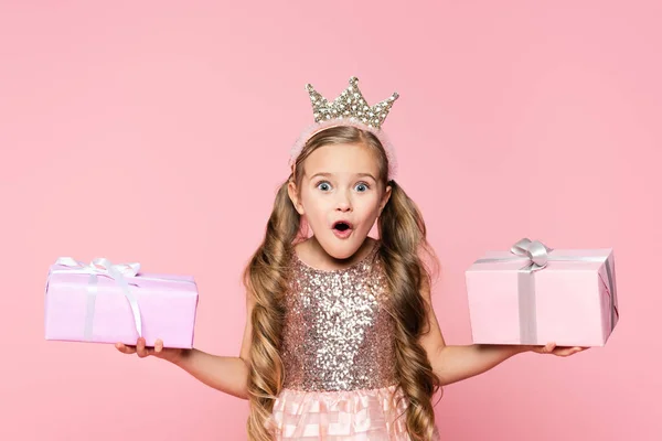 Impactada niña en corona sosteniendo regalos envueltos aislados en rosa - foto de stock