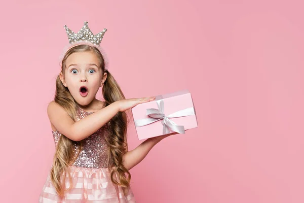 Amazed little girl in crown holding wrapped present isolated on pink — Stock Photo