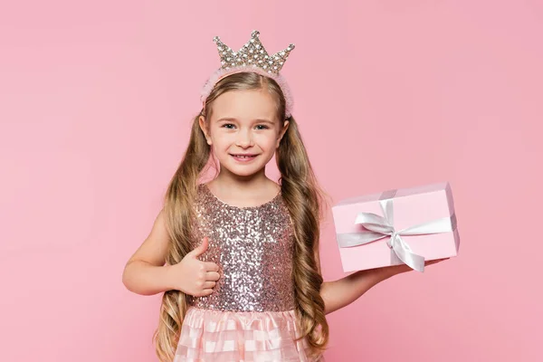 Радісна маленька дівчинка в короні тримає загорнутий подарунок, показуючи великий палець вгору ізольовано на рожевому — стокове фото