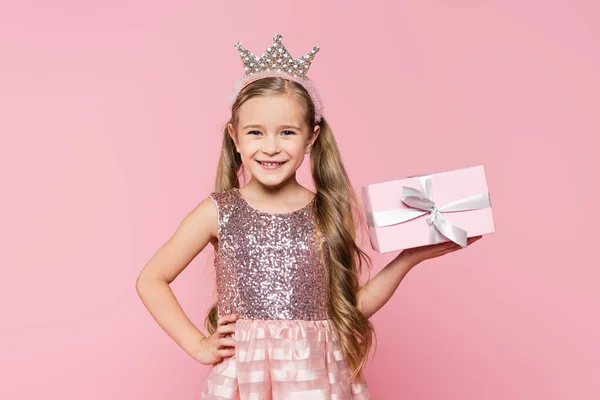 Joyeuse petite fille en couronne tenant enveloppé présent et debout avec la main sur la hanche isolé sur rose — Photo de stock