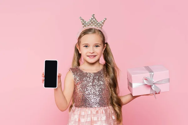 Joyful little girl in crown holding wrapped present and smartphone with blank screen  isolated on pink — Stock Photo