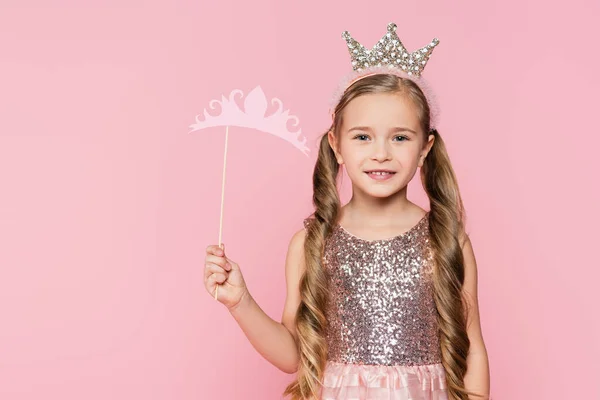 Cheerful little girl in dress holding carton crown on stick isolated on pink — Stock Photo