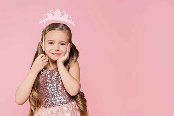 Cheerful little girl in dress holding carton crown on stick above head isolated on pink — Stock Photo