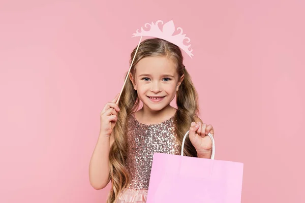 Cheerful little girl in dress holding paper crown on stick above head and shopping bag isolated on pink — Stock Photo