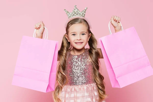 Весела маленька дівчинка в сукні і короні тримає сумки для покупок ізольовані на рожевому — стокове фото