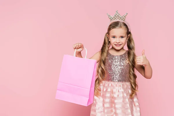 Cheerful little girl in dress and crown holding shopping bag and showing thumb up isolated on pink — Stock Photo