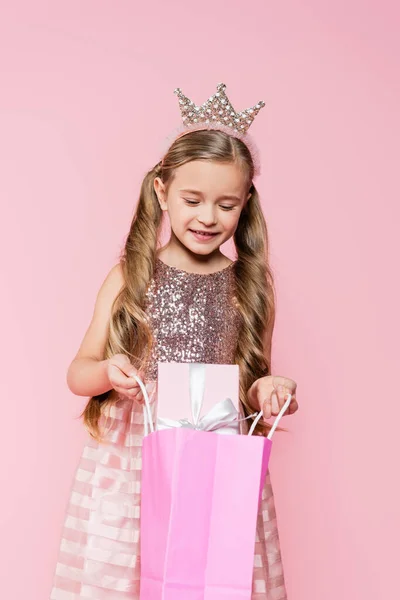 Niña en vestido y corona sosteniendo bolsa de compras con presente aislado en rosa - foto de stock