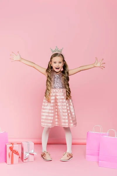 Pleine longueur de petite fille excitée en couronne debout avec les mains tendues près des cadeaux et des sacs à provisions sur rose — Photo de stock